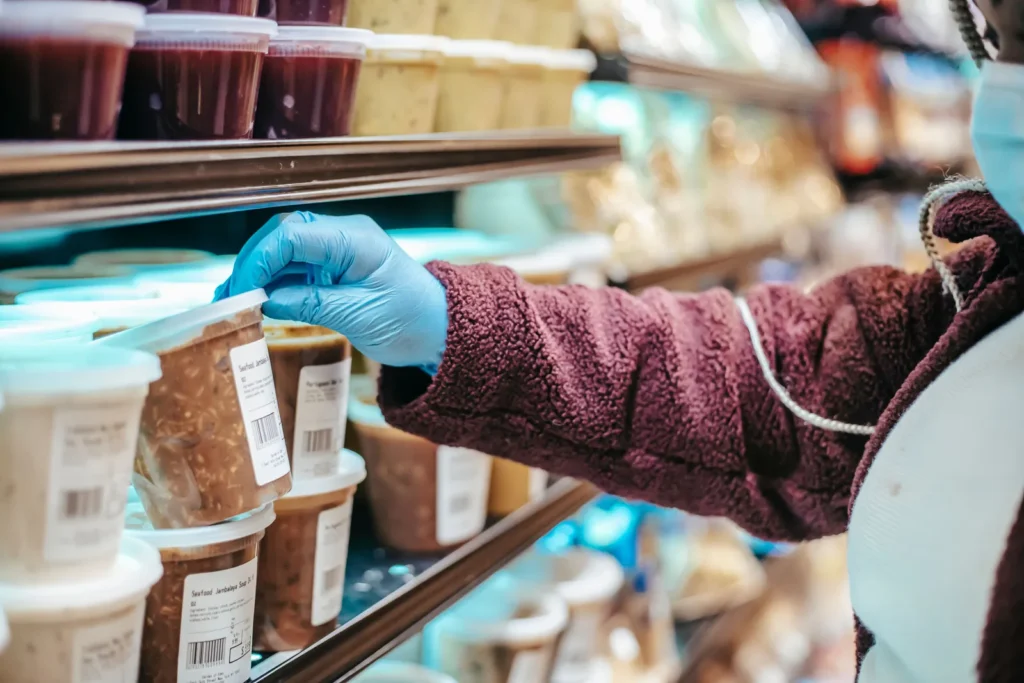 Hand in blauem Handschuh hält eine beschriftete Lebensmitteldose neben Behältern im Ladenregal, Etikettenprüfung bei Lebensmittelallergie in Mekka