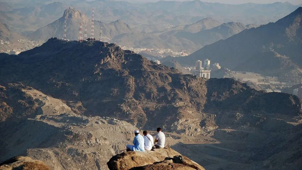Drei Besucher sitzen auf einem Felsvorsprung hoch am Berghang über Mekka – Ausblick bei einem Hira-Besuch mit Kindern und Großeltern