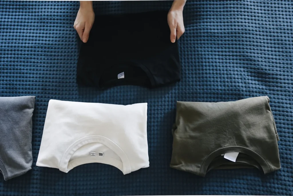 Hands folding a black T-shirt beside folded white, grey and green shirts on a blue bedspread