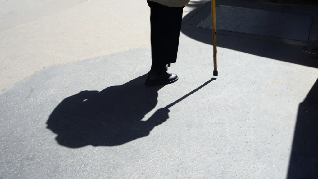 Ombra di una persona anziana che cammina lentamente con il bastone su un marciapiede al sole, durante una ziyarah con genitori anziani