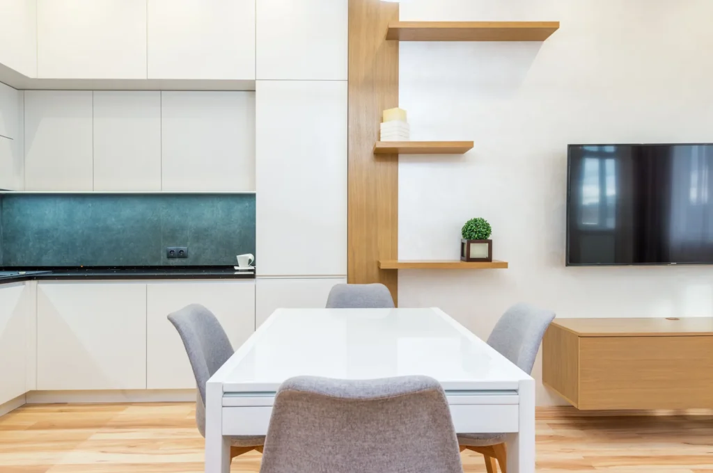 Weiße Küchenschränke neben einem Esstisch mit grauen Stühlen und Holzregalen, allgemeines Bild zur Wahl zwischen Apartment oder Hotel in Mekka
