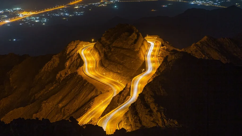 Aerial view of a winding highway lit by car light trails as it climbs through rocky mountains at night
