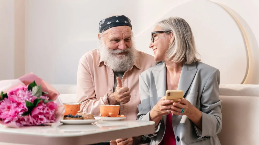 Coppia di anziani sorridenti al tavolino di un caffè mentre uno controlla il telefono, per una ziyarah prenotata da un parente